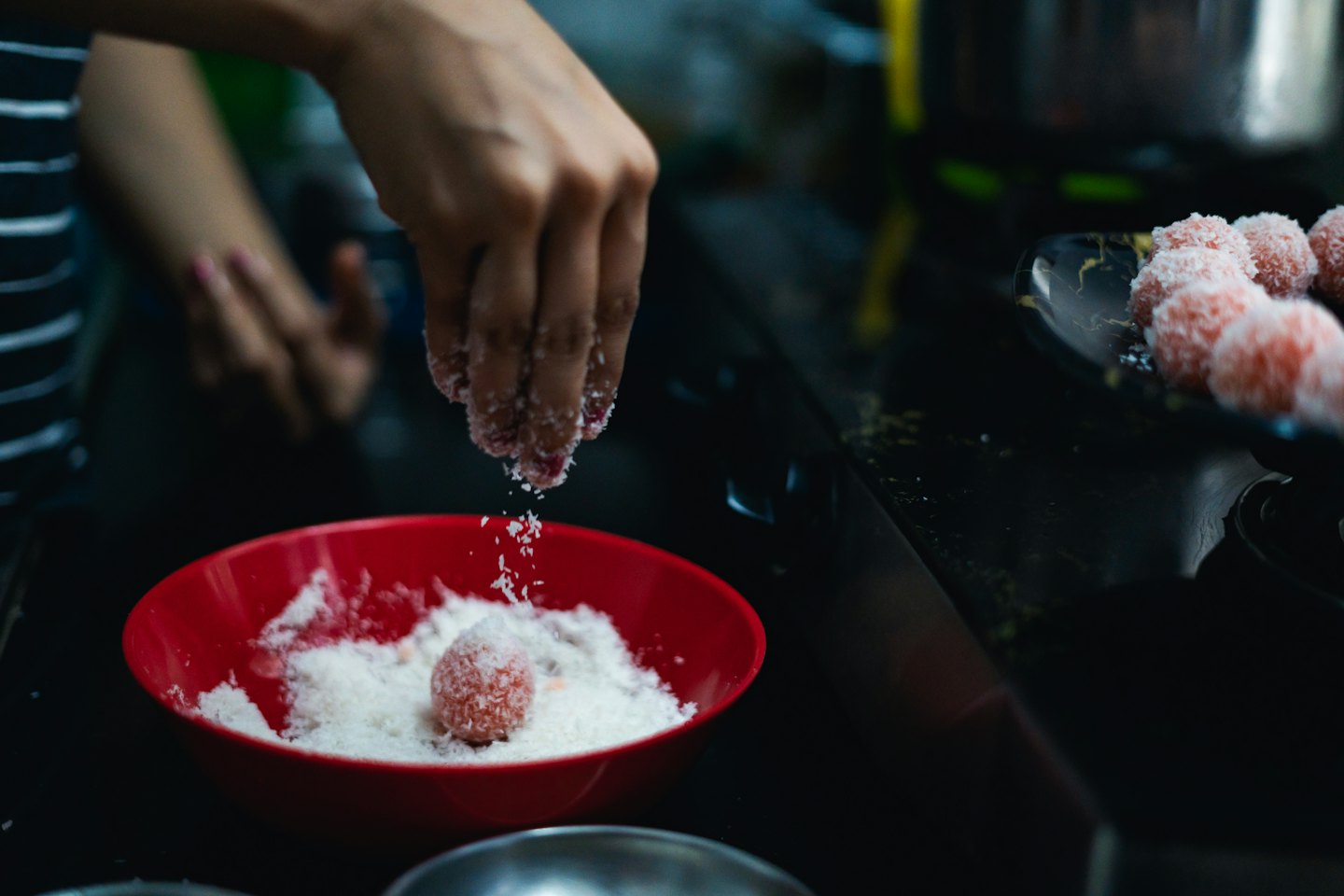 Hands sprinkle coconut over pink sweets in a red bowl.